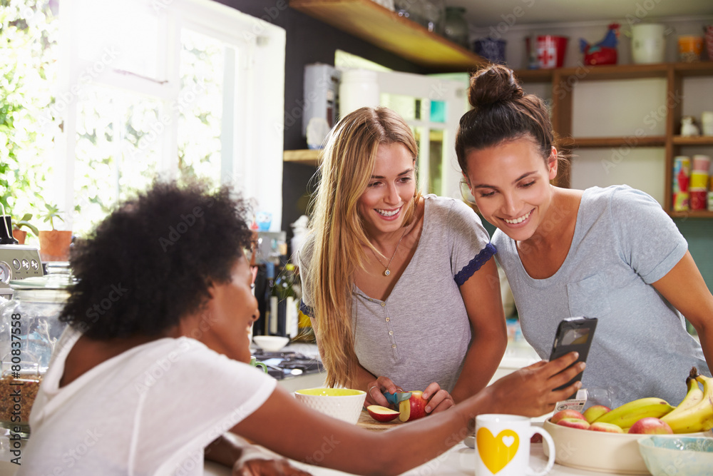 Female Friends Making Breakfast Whilst Checking Mobile Phone Female Friends Making Breakfast Whilst Checking Mobile Phone