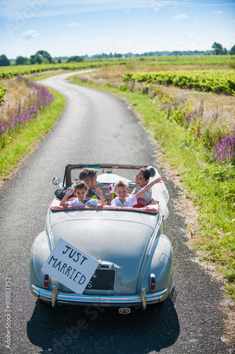 A newlywed couple is driving a retro car with their kids