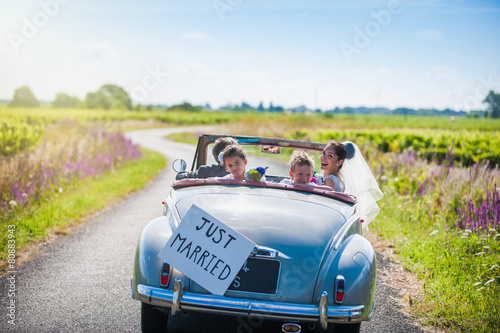 A newlywed couple is driving a retro car with their kids