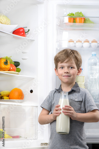 Little cute boy holding bottle of milk near open fridge
