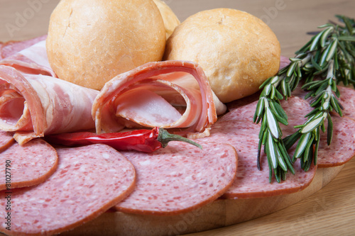 Slices of sausage and bacon on a cutting board with bread rolls,