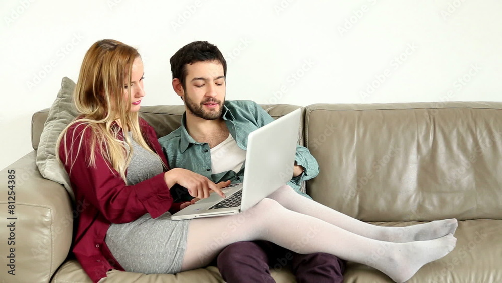 Young couple sitting and talking on couch looking at laptop at home Young couple sitting and talking on couch looking at laptop at home