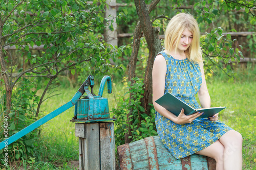 Blonde student girl in garden is reading book with blue cover photo