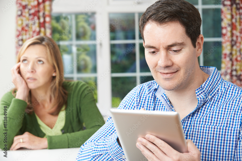 Unhappy Woman Sitting At Table As Partner Uses Digital Tablet Unhappy Woman Sitting At Table As Partner Uses Digital Tablet