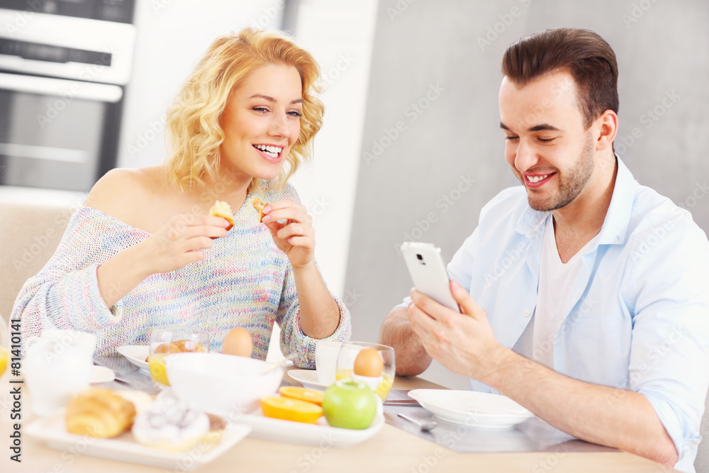 Happy couple eating breakfast and using smart phone