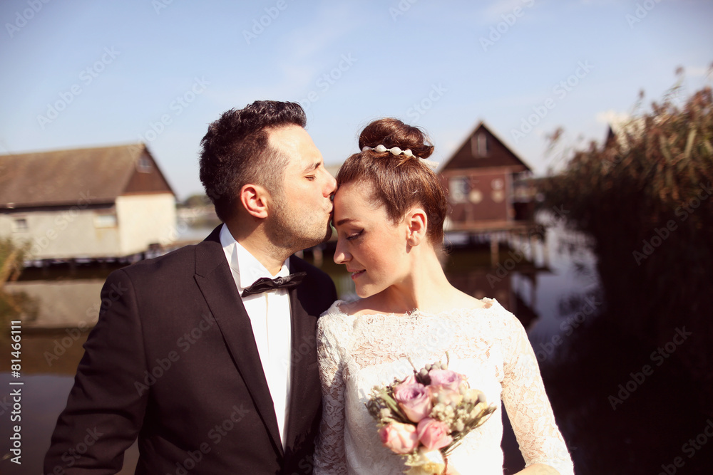 Groom kissing her beautiful bride on bridge near lake houses