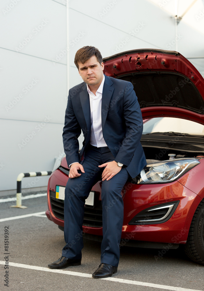 man in suit sitting on broken car with open hood