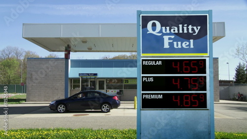 Car drives up to gas pumps at unbranded service station.  Gas prices on sign rise from 3.00 to more than 6.00 (currency not shown so can be used internationally).  Locked off shot.