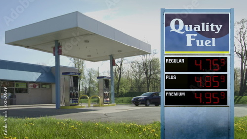 Woman pulls up and gets out of car to pump gas at unbranded gas station.  Prices on sign increase from 2.00 to 13.00.  Currency not specified.  Locked off shot.