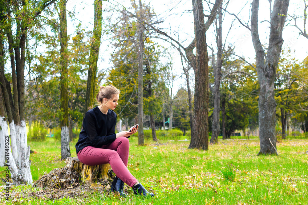 Beautiful girl sitting on a stump in the park with smartphone