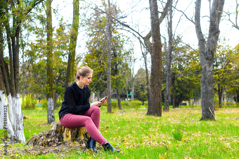 Beautiful girl sitting on a stump in the park with smartphone Beautiful girl sitting on a stump in the park with smartphone
