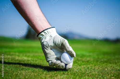 Hand with a glove is placing a tee with golf ball in the ground.