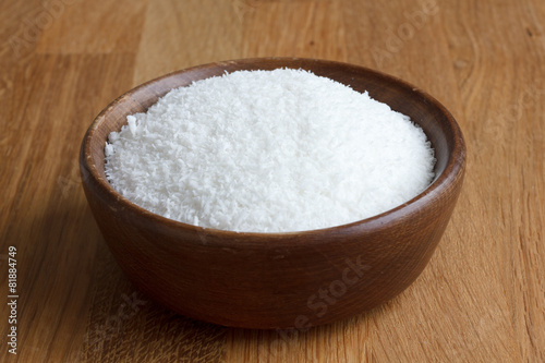 Dark wood bowl of dessicated coconut on wood table.