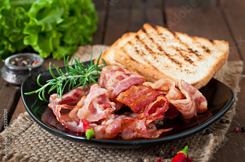 Fried bacon and toast on a black plate on a wooden background © timolina