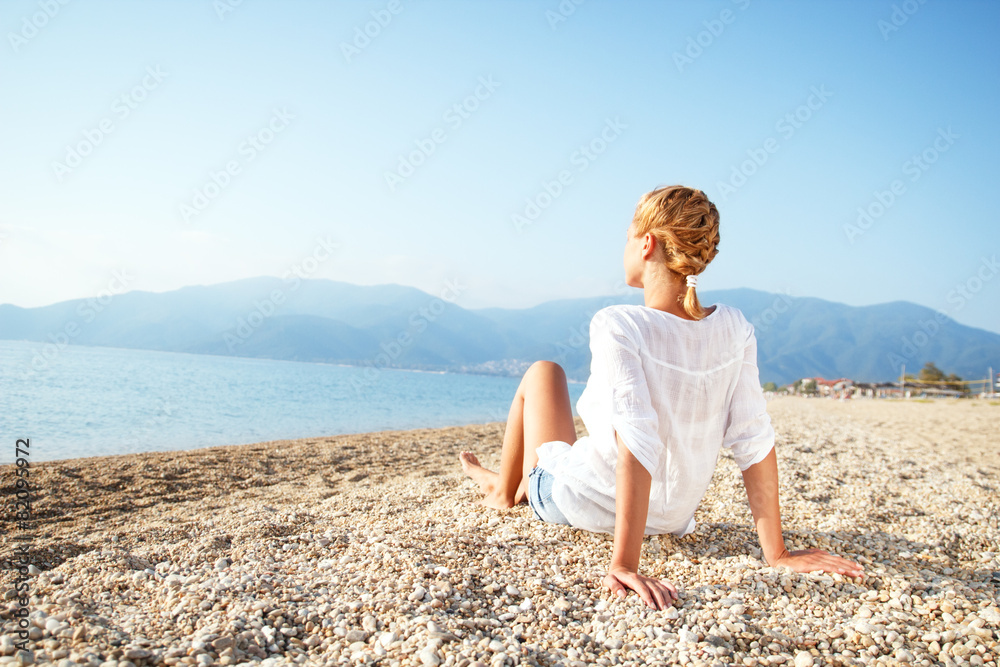 Rear view of young woman sitting on the beach and looking on horizon.
