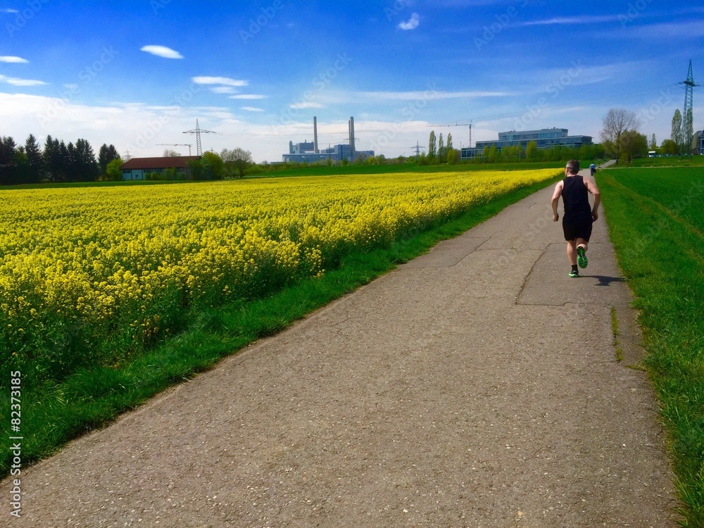 Obraz premium Jogger am Rapsfeld läuft Richtung Heizkraftwerk