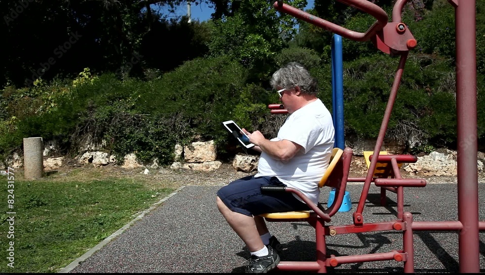 A big lazy man with sunglasses uses a tablet in outdoor gym