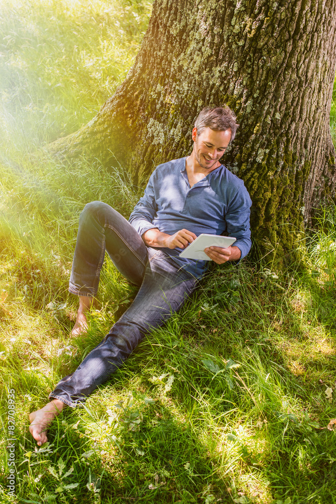 A gray-haired man sitting against a tree looking at his tablet