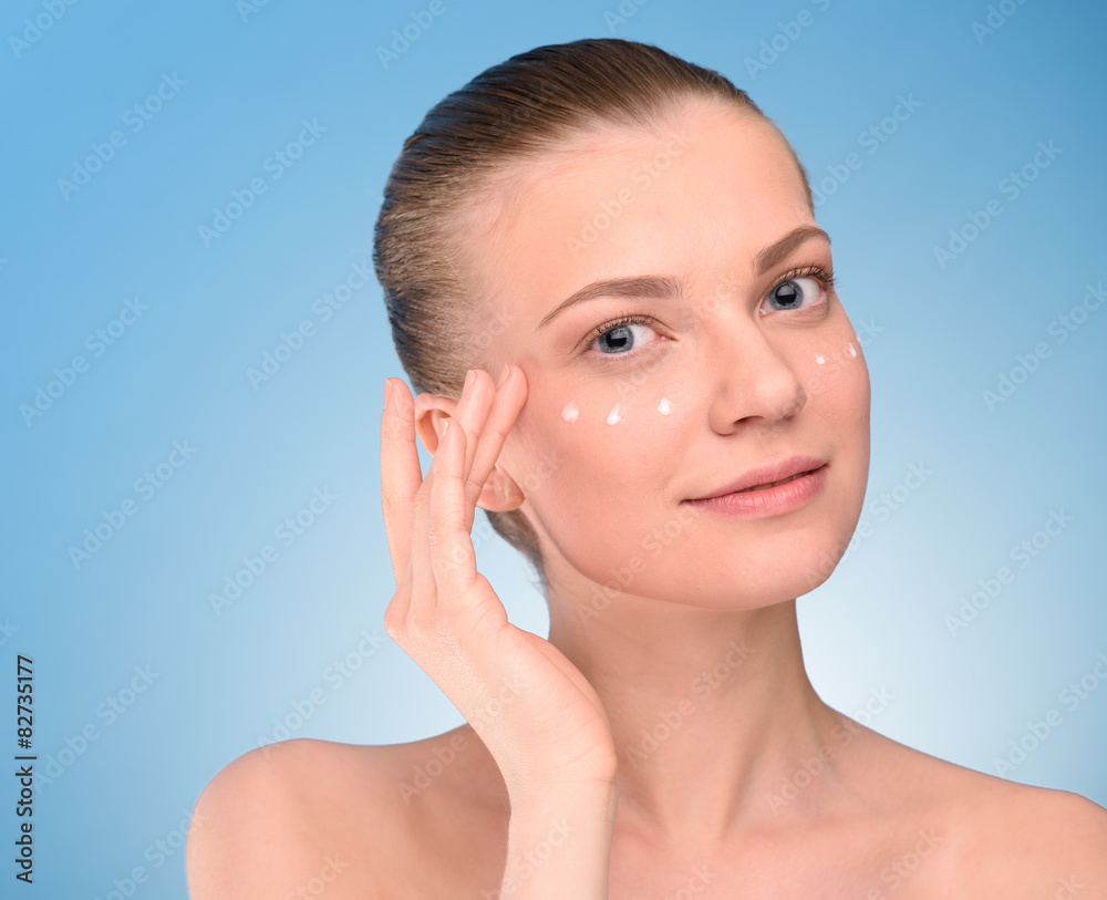 Woman applying lifting cream on face, over blue background. 