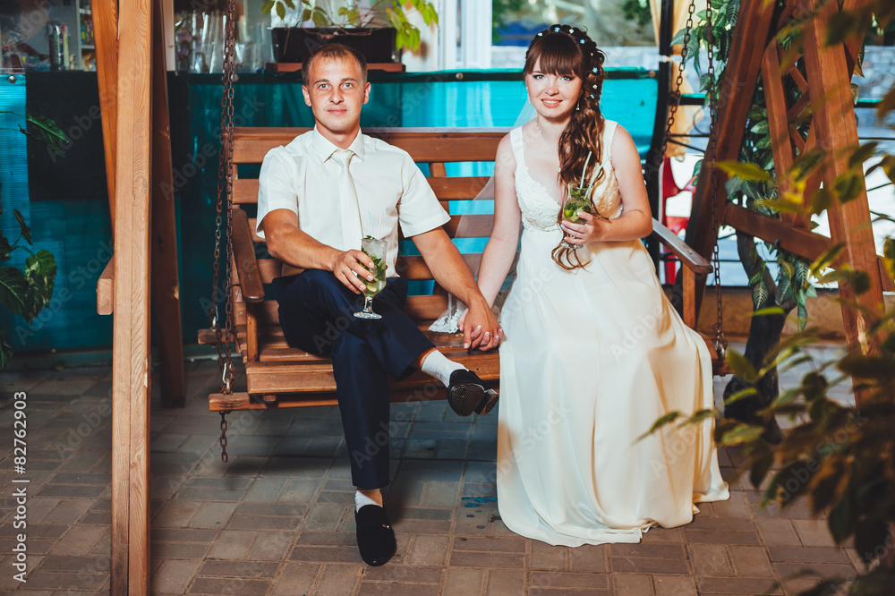 Obraz premium happy bride and groom drink mojito, swinging on a swing