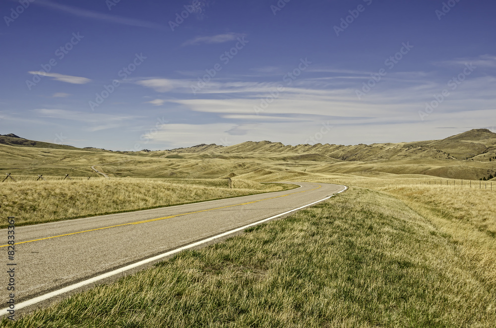 Road with a curve and no traffic that can be seen running through rural Montana with fields and mountains
