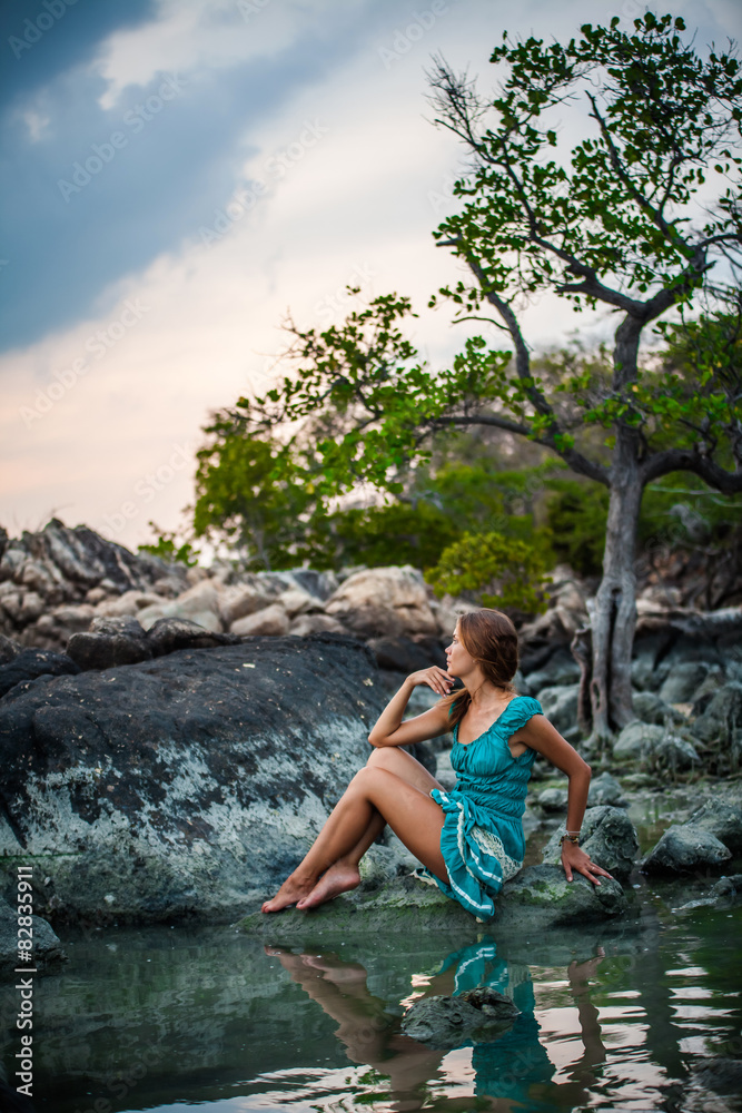 Fototapeta premium Young beautiful woman in long turquoise dress sitting on a stone