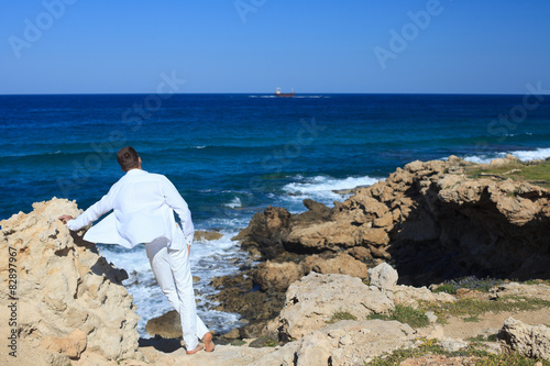 guy in the white suit leaning on a rock and looking at sea.