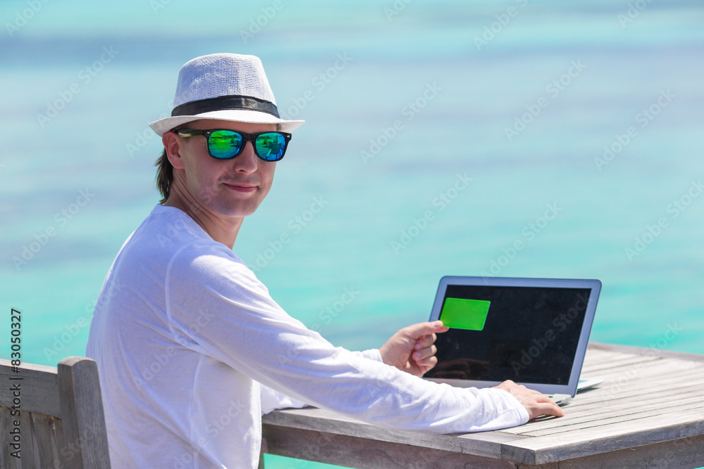 Young man working on laptop with credit card at tropical beach