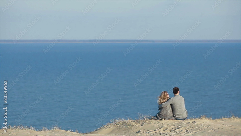 Young couple in love sit embracing on the top of a natural dune hill enjoying the fascinating vast view of blue calm sea Young couple in love sit embracing on the top of a natural dune hill enjoying the fascinating vast view of blue calm sea