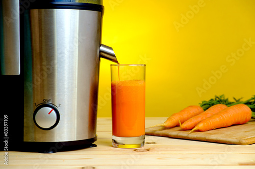 Juicer and carrot juice in glass on yellow background