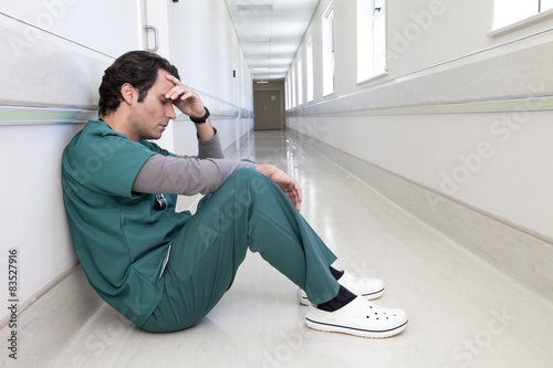 Tired doctor sitting on the floor in a hospital corridor with his hand on his head photo