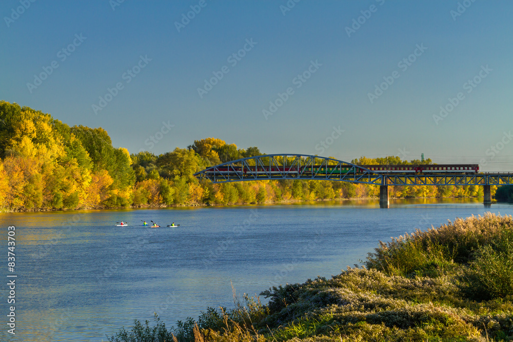 Obraz premium Autumn at river with kayaks and train crossing bridge.