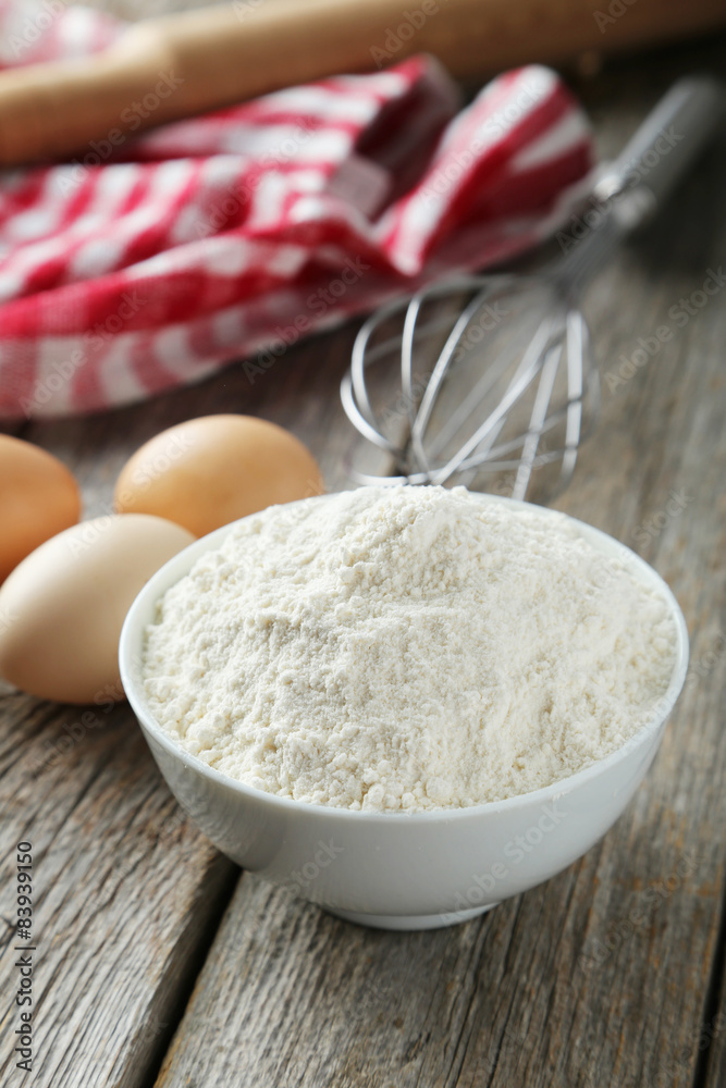 Bowl of wheat flour with eggs and whisk on grey wooden backgroun