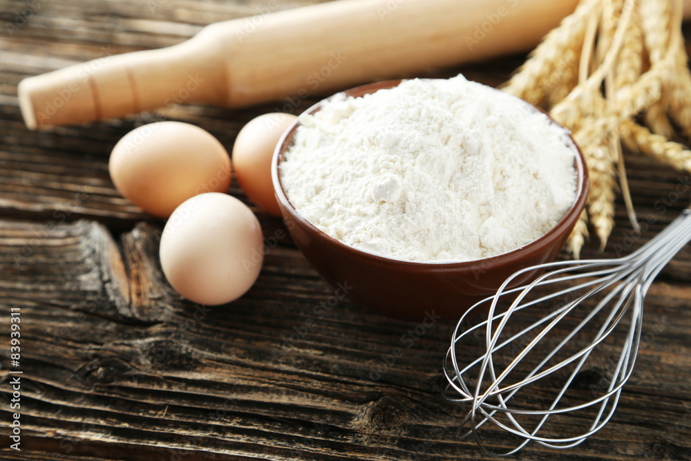 Bowl of wheat flour with eggs and whisk on brown wooden backgrou Bowl of wheat flour with eggs and whisk on brown wooden backgrou
