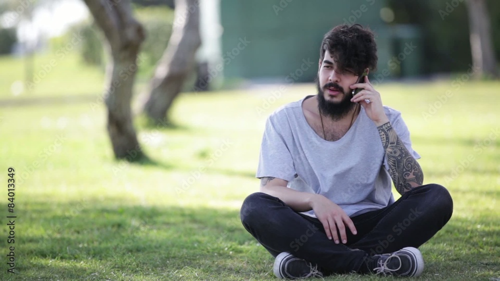 young hipster guy sitting on grass talking on his smart phone
