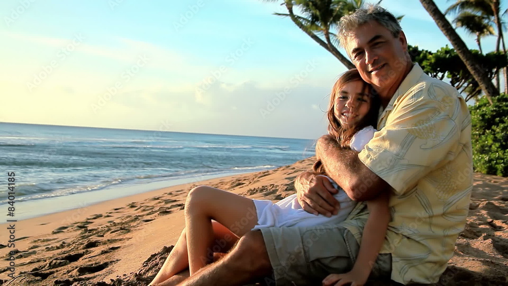 A little girl sitting in her father's lap on the beach in Hawaii.