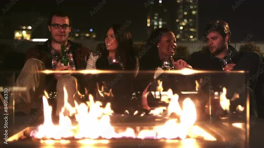 Two couples hanging out on a rooftop bar at night sit in front of a fire pit and drink Two couples hanging out on a rooftop bar at night sit in front of a fire pit and drink