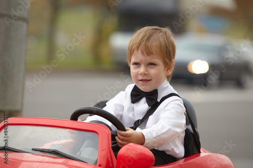 Photography year-old boy in a white shirt in a red toy car in the street