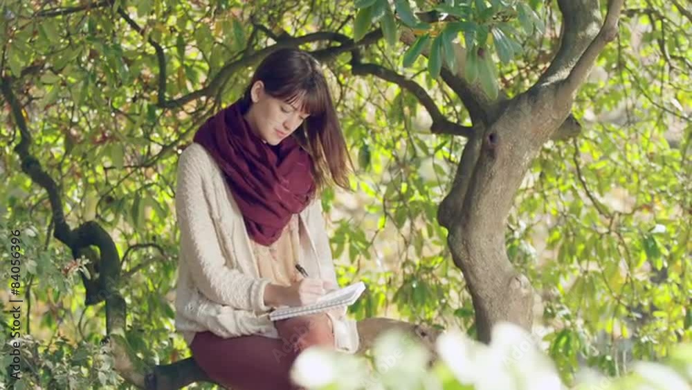 A young woman sitting in a tree writes in her notebook and smiles A young woman sitting in a tree writes in her notebook and smiles