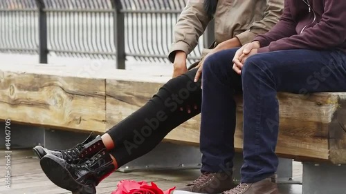 A couple sits on a bench in a park in front of the brooklyn bridge on an overcast day, close up in slow motion
