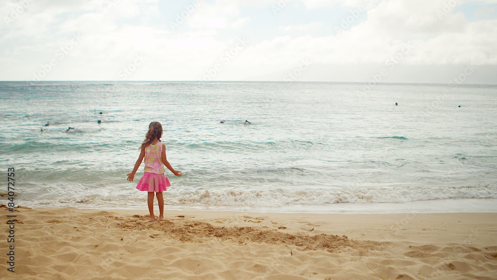 A young girl stands on the beach and stares at the waves hitting the shore A young girl stands on the beach and stares at the waves hitting the shore