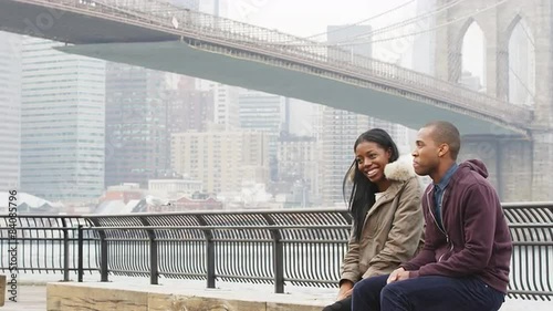 A couple sits on a bench in a park in front of the brooklyn bridge on an overcast day