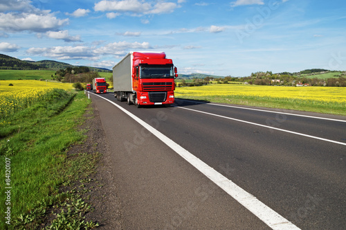 Obraz na płótnie Three red trucks on the road between rapeseed field w salonie