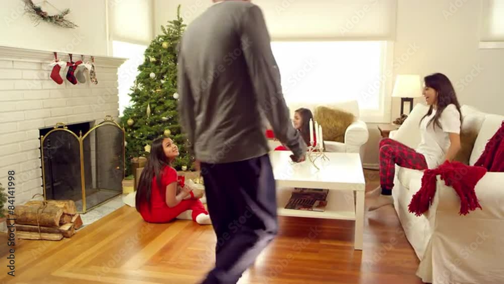 A father walks in to see his two daughters sitting under the tree and opening their christmas presents A father walks in to see his two daughters sitting under the tree and opening their christmas presents