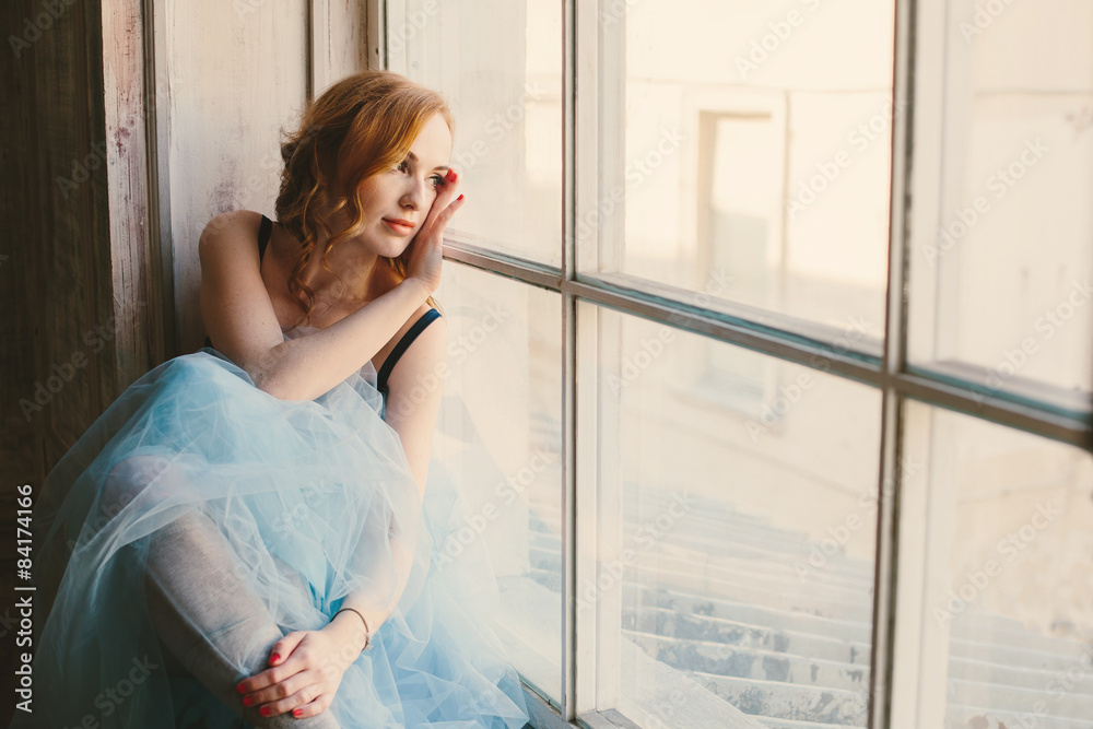 Toned portrait of a young woman with red hair and blue fluffy skirt near the big window