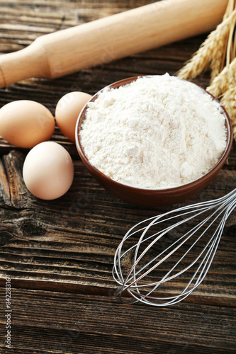 Bowl of wheat flour with eggs and whisk on brown wooden backgrou