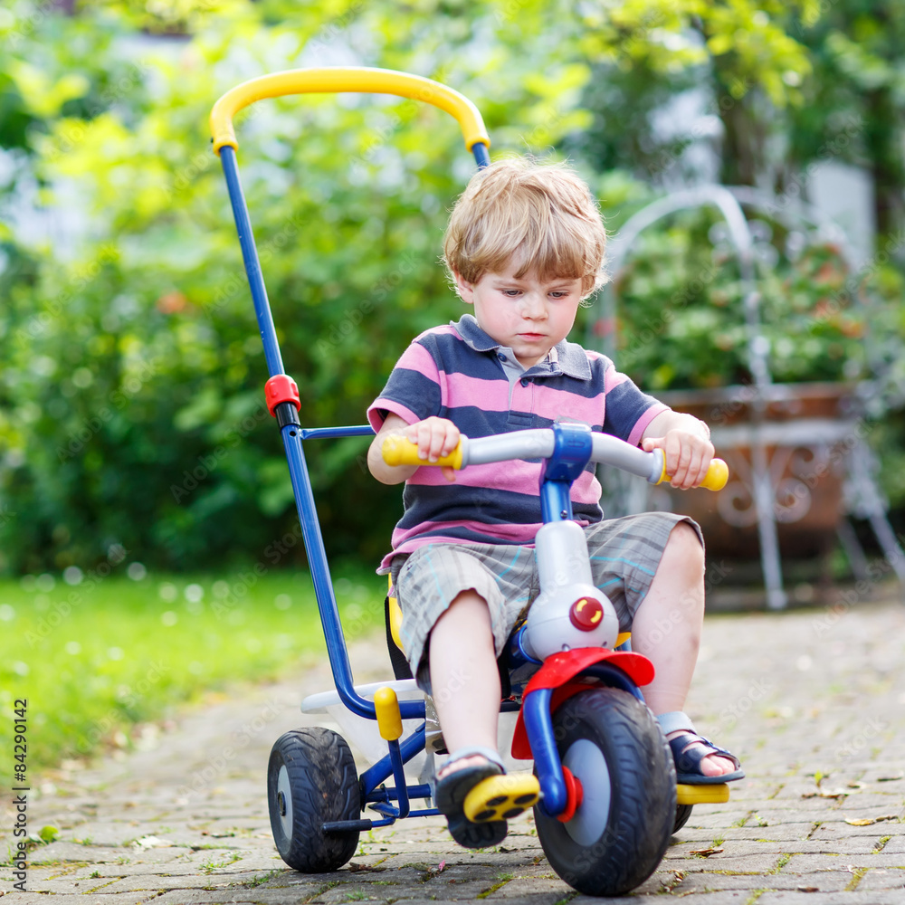 Active blond kid boy driving tricycle or bicycle in domestic gar Active blond kid boy driving tricycle or bicycle in domestic gar