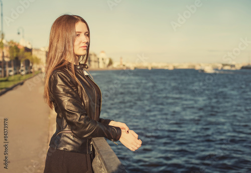 Pretty young woman student with long hair in leather jacket thinking near city river