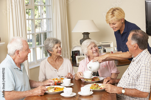 Group Of Senior Couples Enjoying Meal Together In Care Home With Home Help