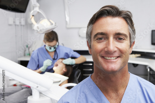 Portrait Of Dental Nurse With Dentist Examining Patient In Background